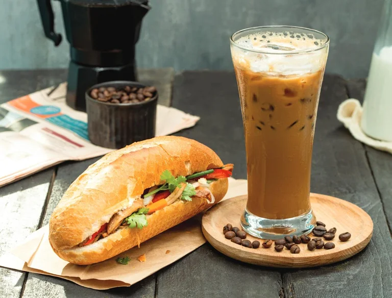 A banh mi sandwich and a glass of Vietnamese iced milk coffee on a wooden table
