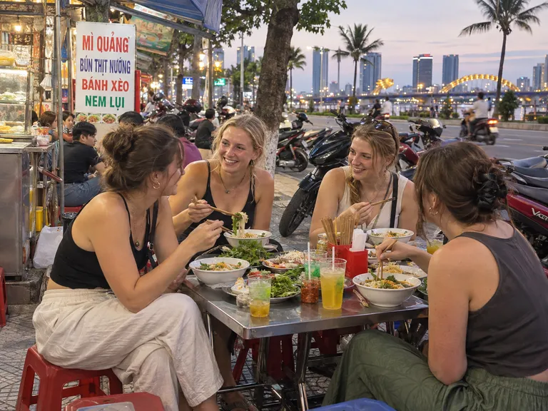 Four friends sharing a street-food dinner with the Dragon Bridge lit up behind them