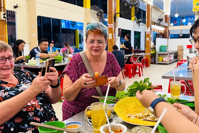 Women photographing banh xeo and fresh herbs at a bustling food hall