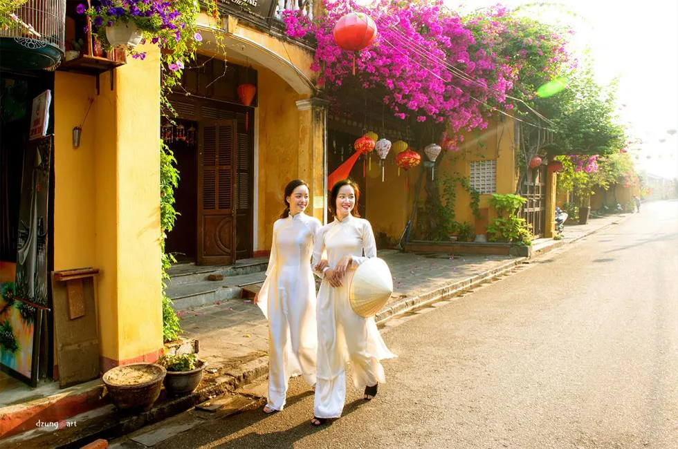Two friends in white ao dai strolling beneath bougainvillea on a sunlit Hoi An street