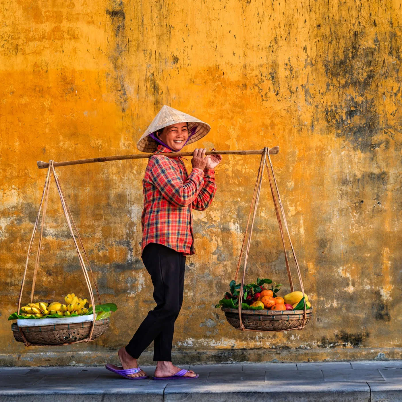 A smiling fruit vendor with a shoulder pole of baskets before a mustard-yellow Hoi An wall