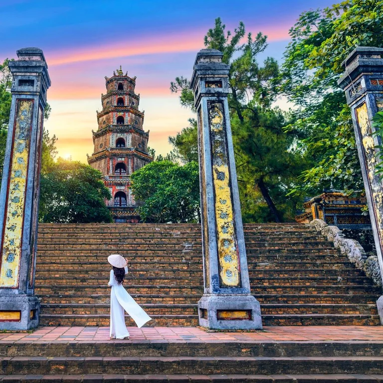 A woman in a white ao dai on the stone steps below Thien Mu Pagoda in Hue at sunset