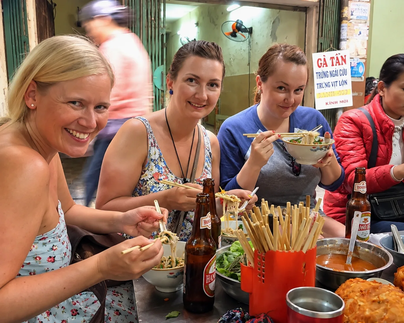 Four smiling friends sharing noodles at a street stall at night
