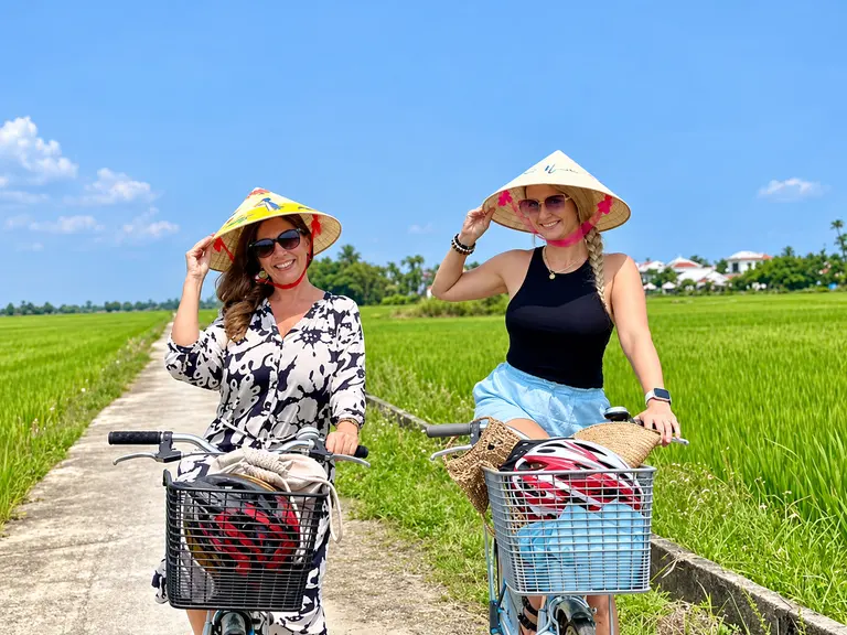 Two friends in conical hats cycling through green rice fields