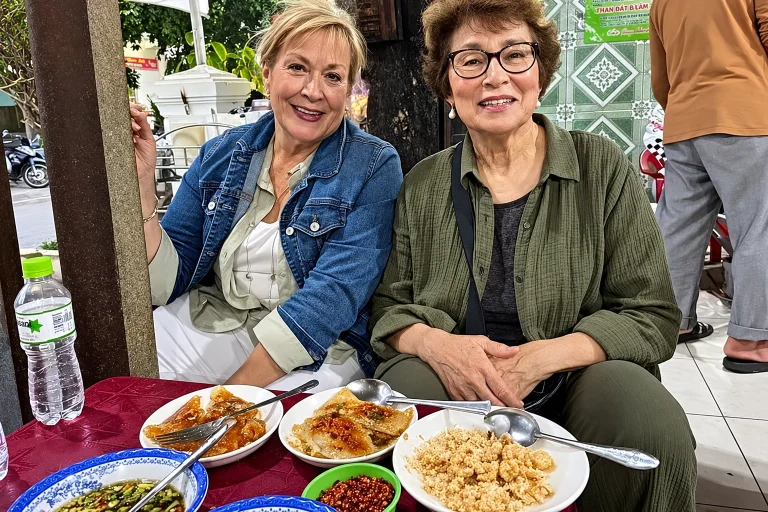 Two women smiling over a table of Vietnamese street-food dishes