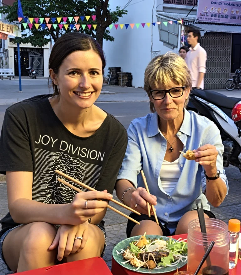 Two women trying street food with chopsticks on a city sidewalk